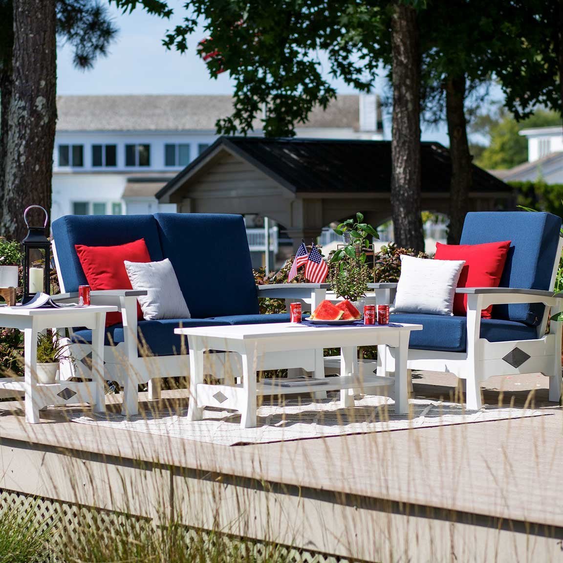White poly loveseat and armchair with navy cushions, red pillows and white tables on a deck near a lake house