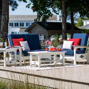 White poly loveseat and armchair with navy cushions, red pillows and white tables on a deck near a lake house