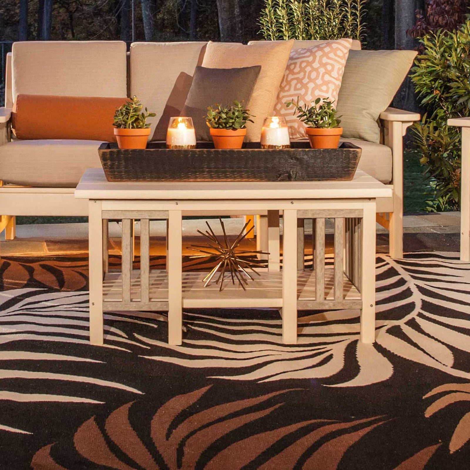 Ivory poly Mission coffee table with slatted sides holding candles and potted plants beside a sofa at dusk