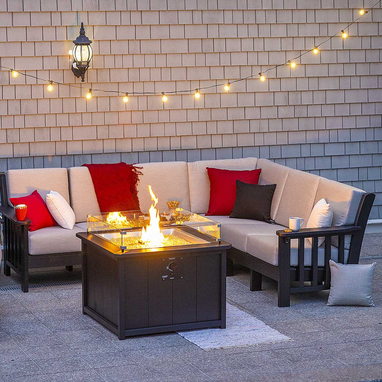 Brown poly L sectional with beige cushions and red pillows around a lit fire table under string lights