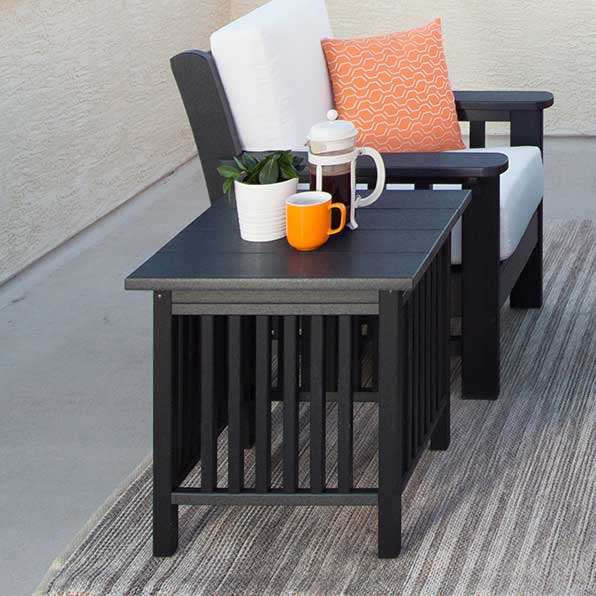 Black poly Mission side table with slatted sides holding coffee and a plant beside a cushioned chair