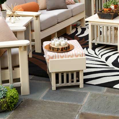 Ivory poly ottoman with patterned cushion beside a matching sofa and side tables on a flagstone patio