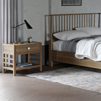 Open one drawer nightstand in light oak with a lower shelf and ladder sides, beside a spindle bed with a lamp on top