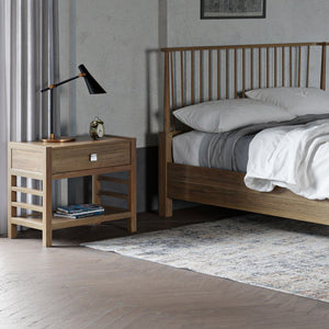 Open one drawer nightstand in light oak with a lower shelf and ladder sides, beside a spindle bed with a lamp on top