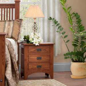 Mission oak three-drawer nightstand with black bail pulls beside a slatted bed, topped with a lamp and lilies