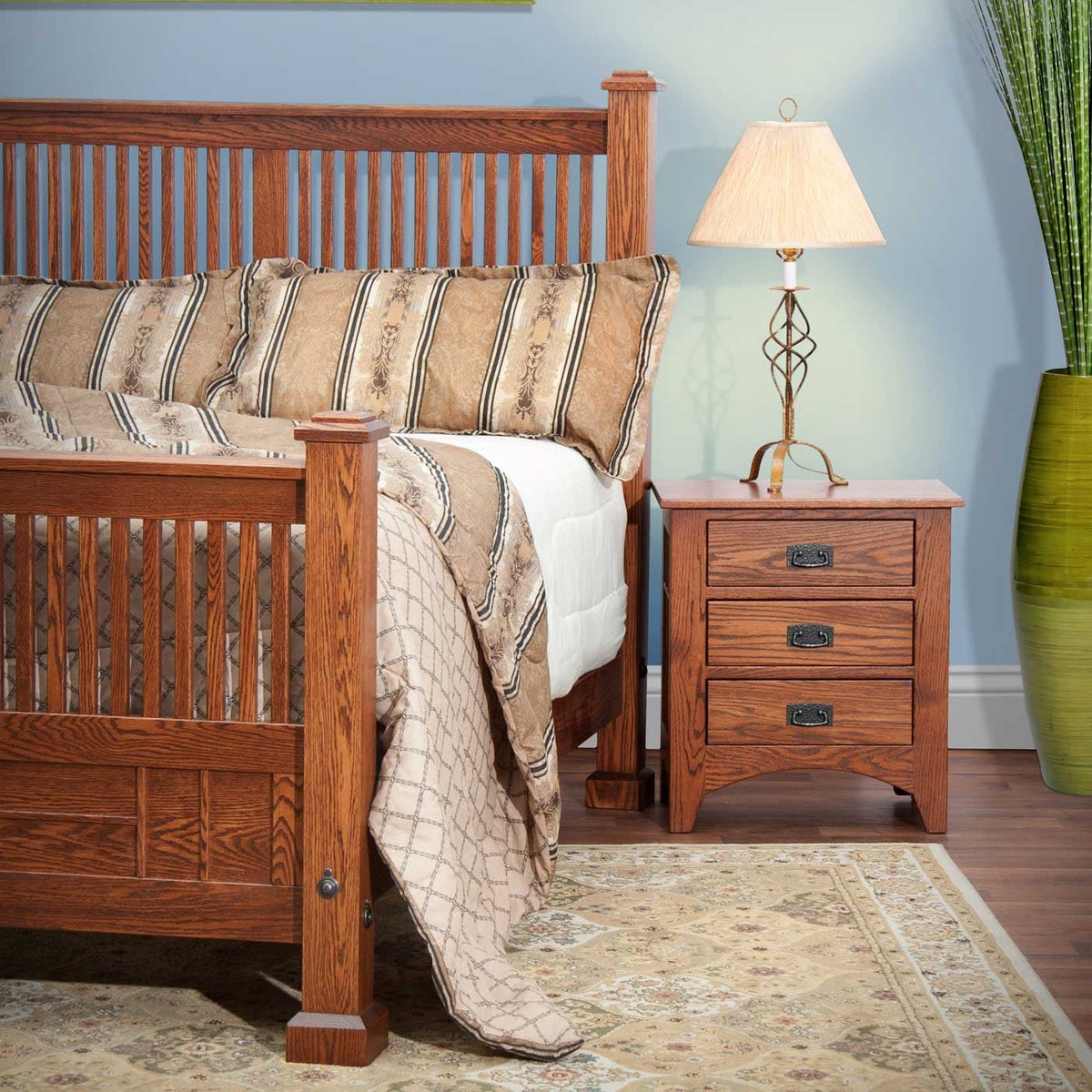 Slatted oak Mission bed with striped bedding next to a three-drawer nightstand and lamp in a blue bedroom