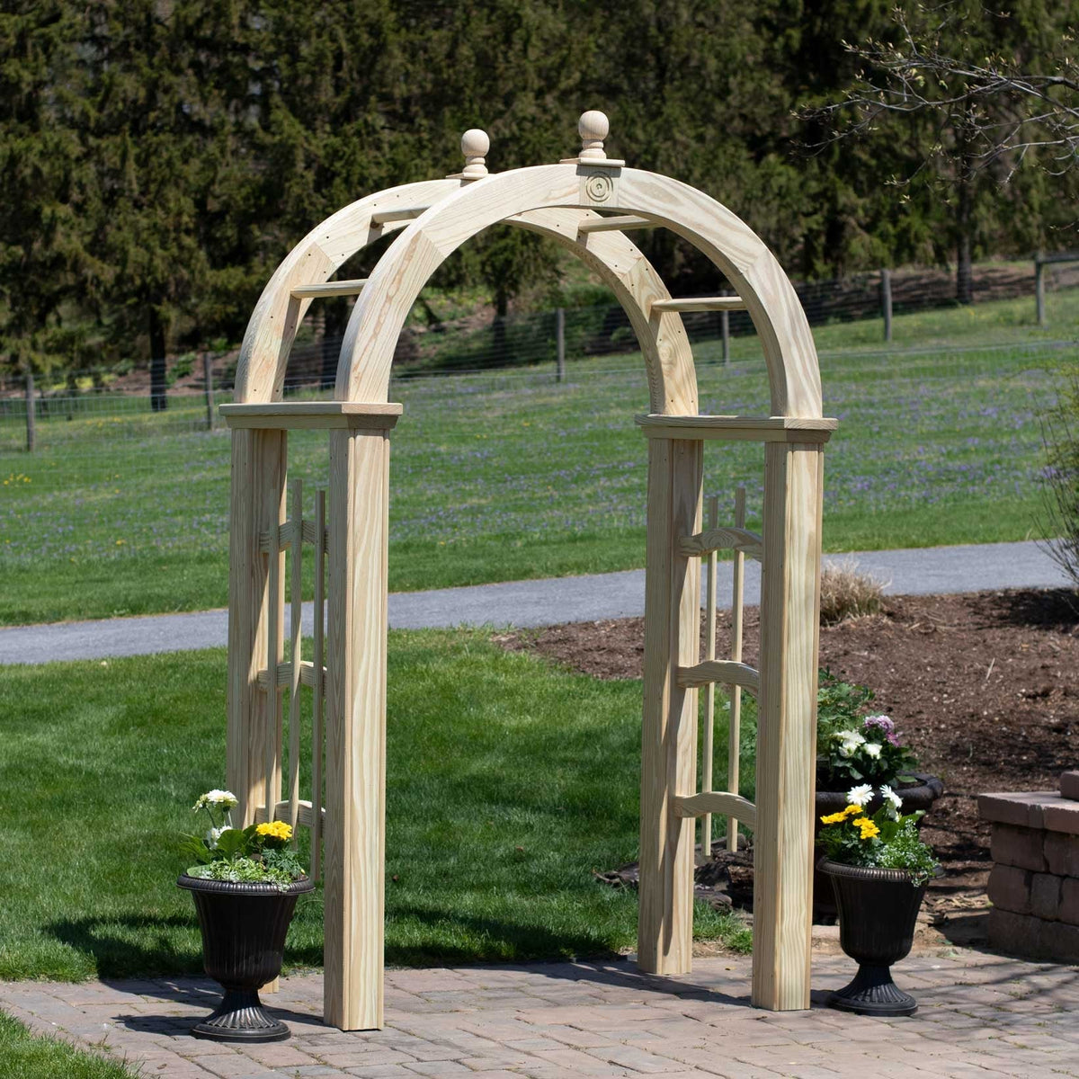 Natural wood arched garden arbor with ball finials flanked by urn planters on a brick patio by a lawn
