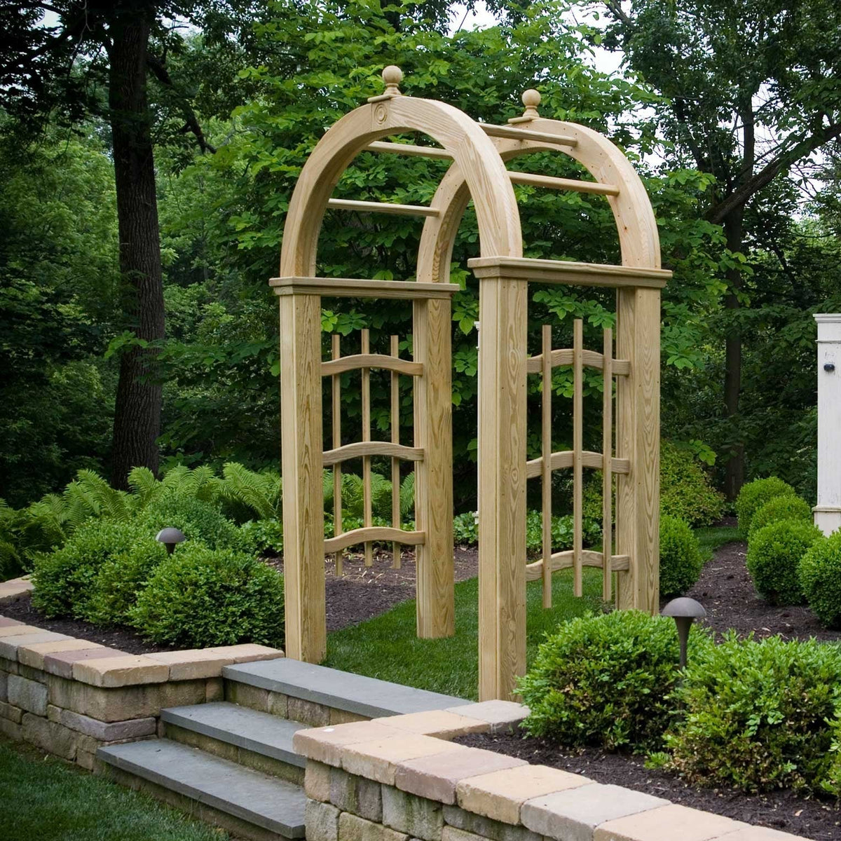 Light wood rounded arch arbor with curved trellis panels set at stone steps in a boxwood and fern garden