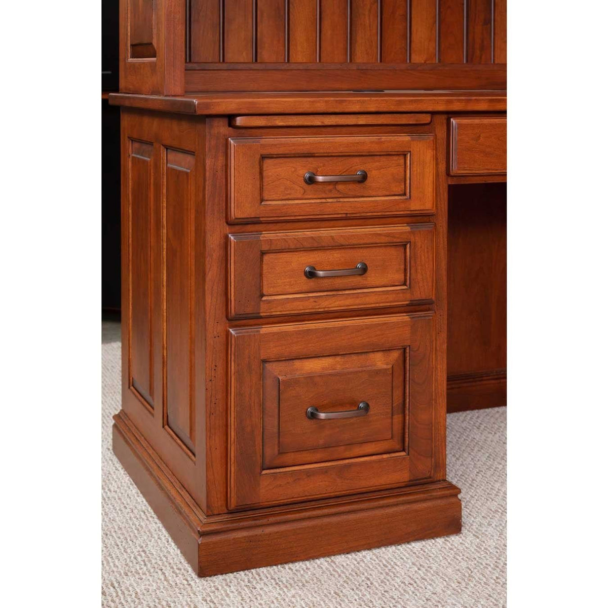 Corner desk pedestal detail with three raised panel drawers, dark bronze pulls and a molded plinth base