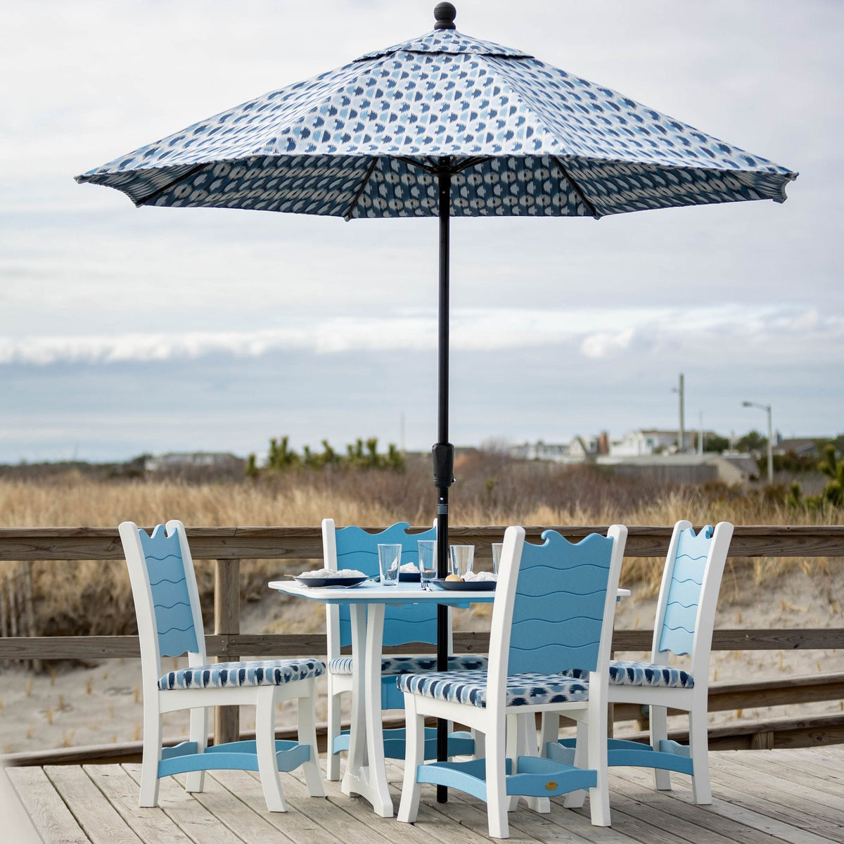 White and blue wave poly dining set for four under a patterned market umbrella on a boardwalk overlooking dunes