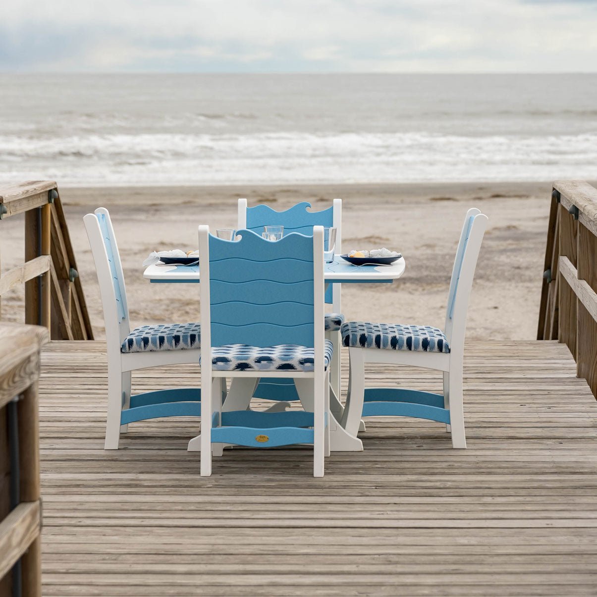 Square wave back poly dining table and four chairs with blue tie dye cushions on a beach boardwalk facing the surf