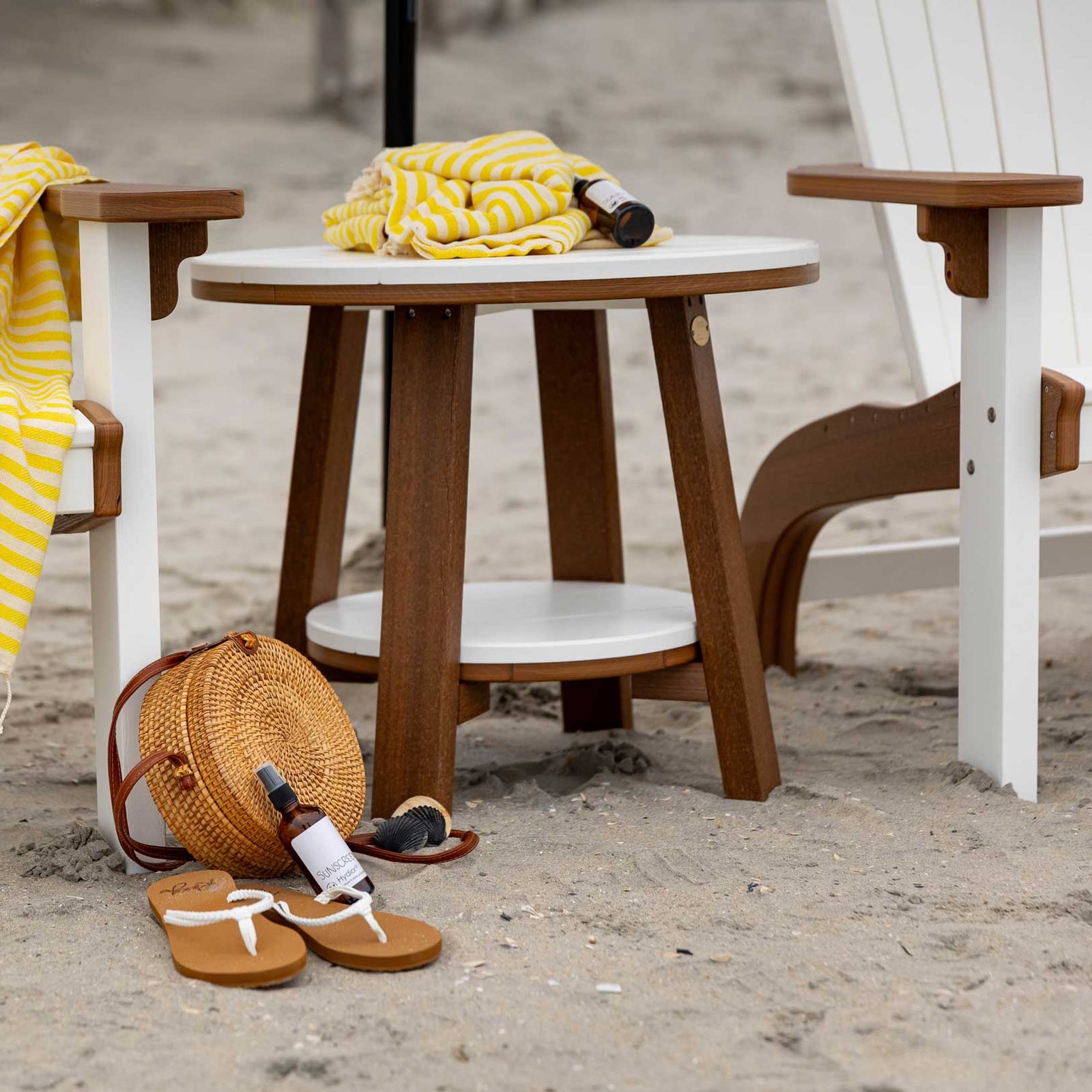 Round white and brown poly bistro table with a lower shelf between two Adirondack chairs on the beach