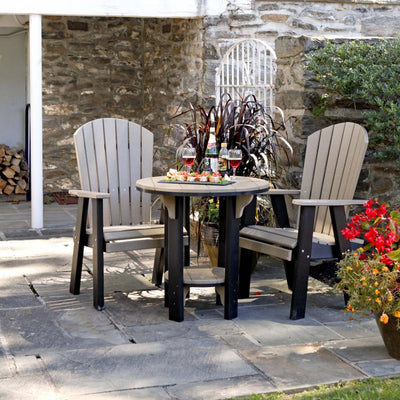 Weatherwood and black poly 30 inch round patio pub table with two tall fan back Adirondack chairs on a stone patio