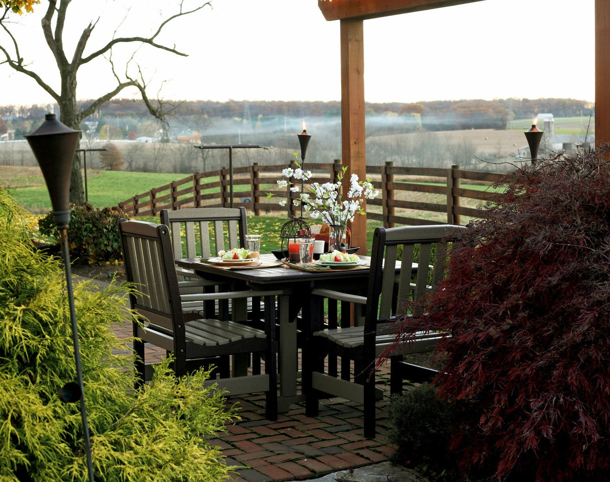 Dark and gray poly English Garden patio table with four slat back chairs on a brick patio with torches at dusk