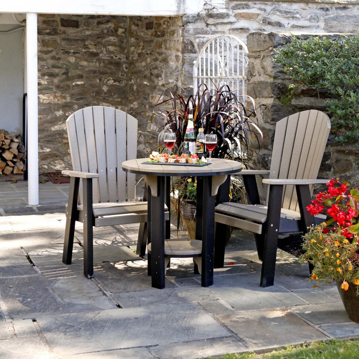 Weatherwood and black poly 30 inch round patio pub table with two tall fan back Adirondack chairs on a stone patio