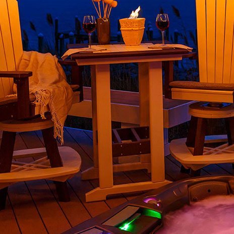 Square poly patio bar table lit at dusk on a deck with swivel Adirondack chairs, wine glasses and a fire pot