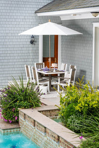 White and driftwood poly patio dining set for six under a white umbrella on a poolside patio with flower beds