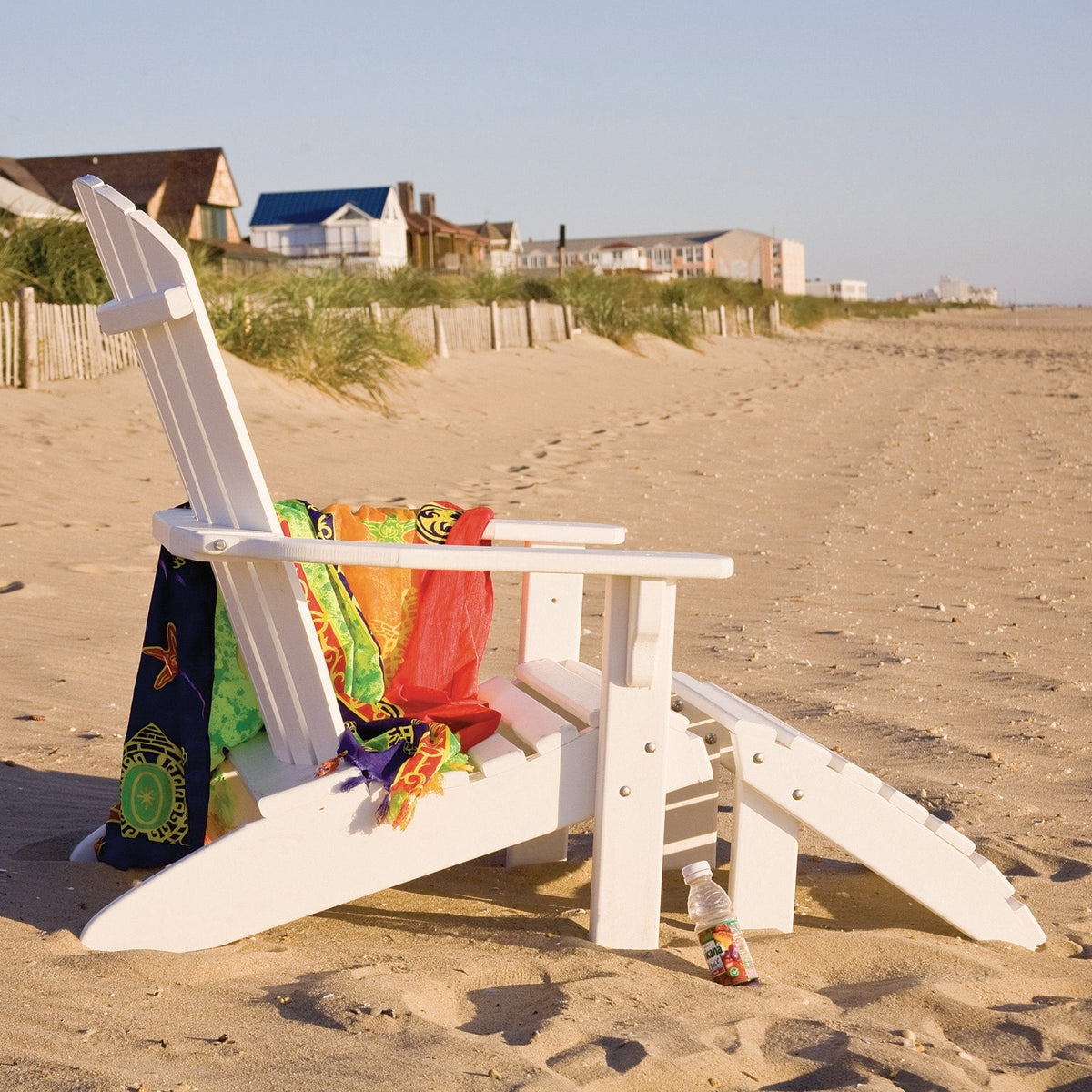 White poly Adirondack chair with slat back, ottoman and beach towels facing dunes and shore houses