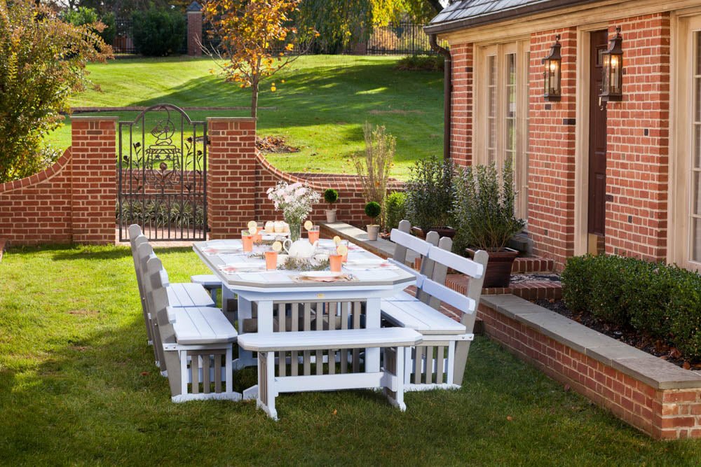 White poly Mission table with four benches set for a party on a lawn beside a brick house and garden gate