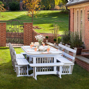 White and driftwood gray poly patio dining table with four slatted benches set for a meal on a lawn beside a brick garden wall