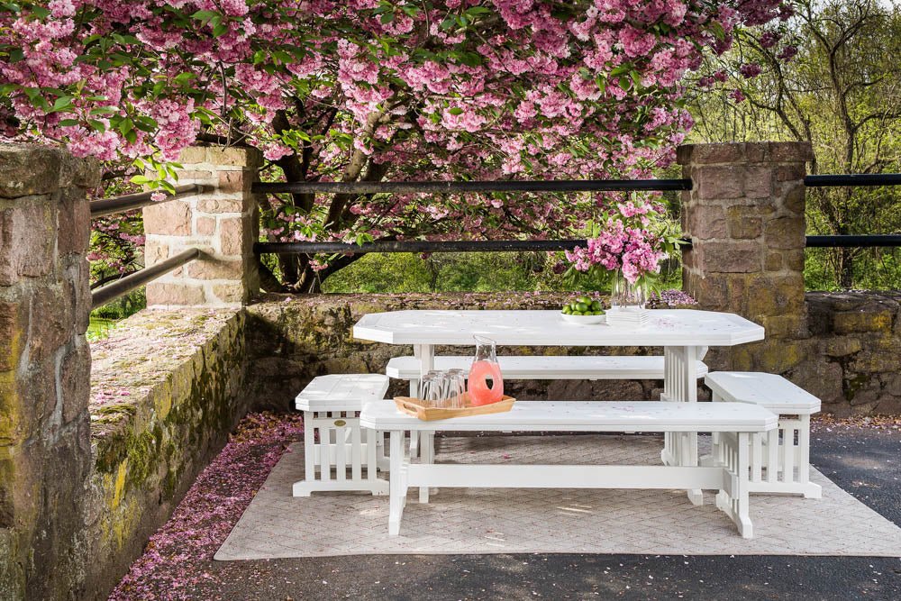 All-white poly patio table with bordered clipped-corner top and three benches on a stone patio under blooming cherry trees