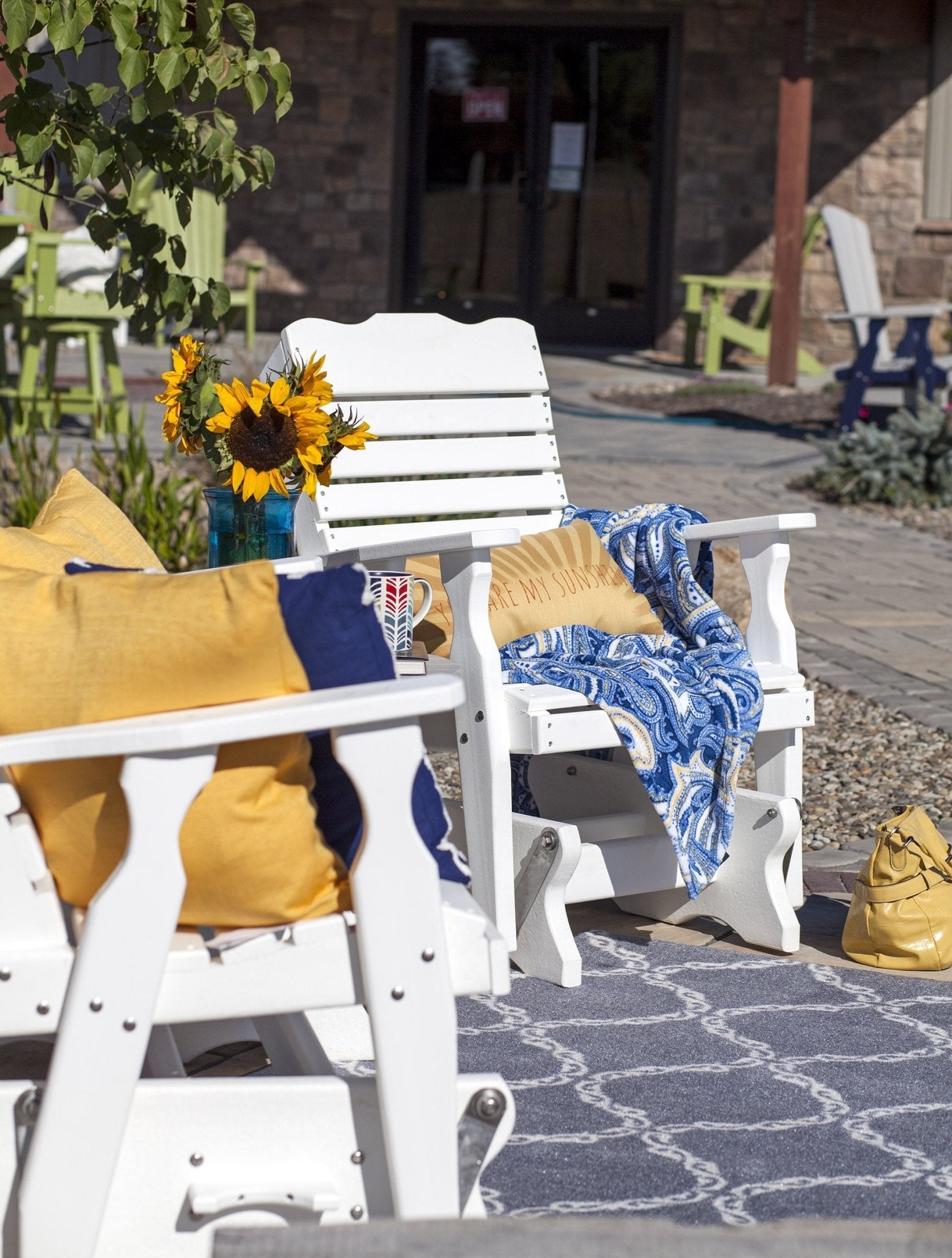 White poly curve back bench glider with pillows and a small round side table on a paver patio by flowering shrubs