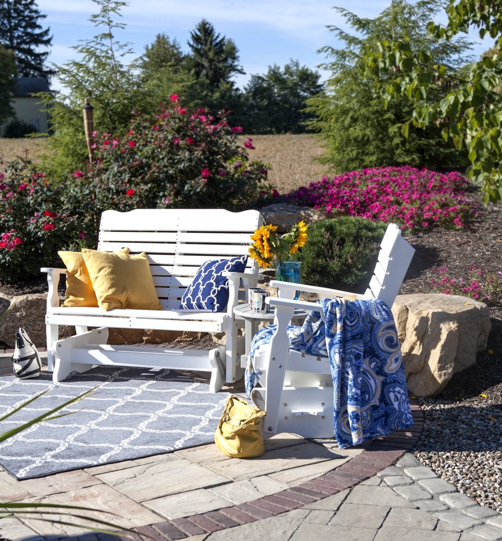White poly curve back bench glider with pillows and a small round side table on a paver patio by flowering shrubs