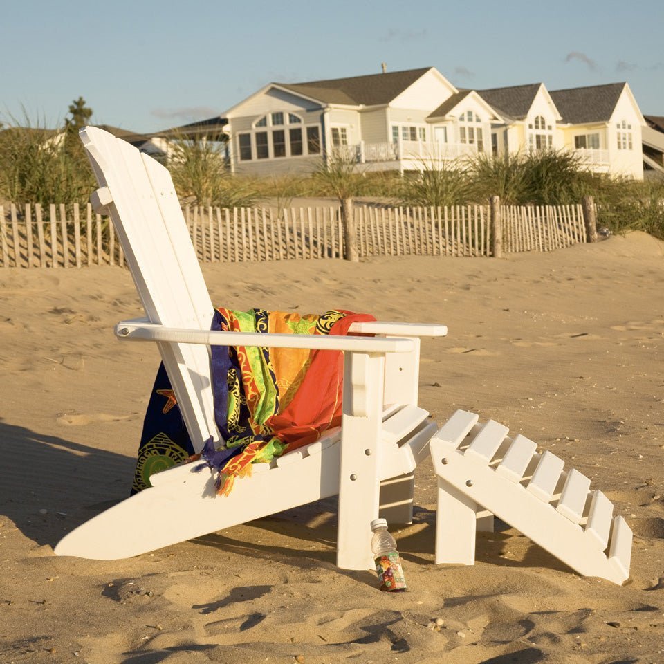 White poly fanback Adirondack chair and matching footrest with towels on the beach near a shingled cottage