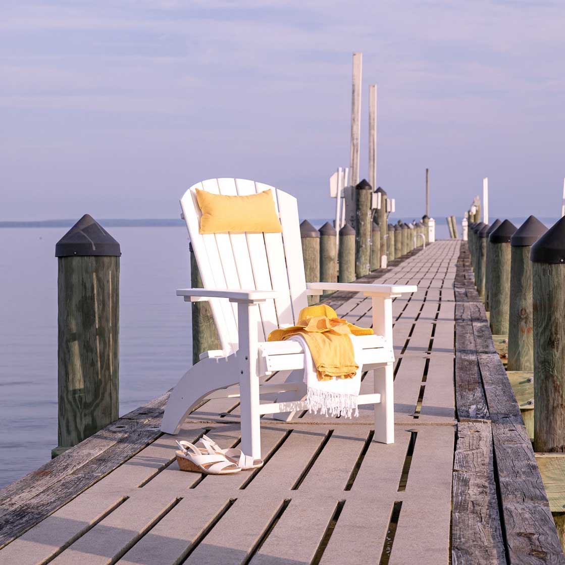 White poly grandpa high Adirondack chair with a yellow pillow and throw on a weathered pier over the bay