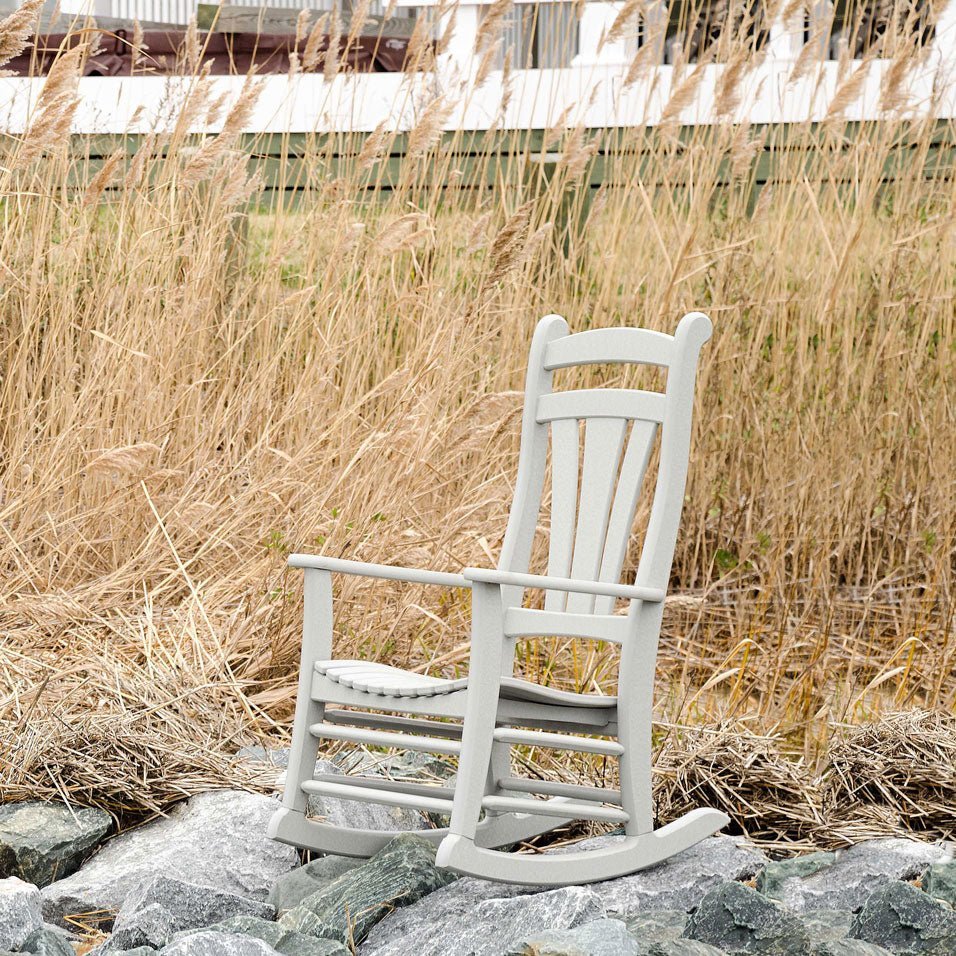 Light gray poly rocking chair with a fanned slat back and scalloped seat set on rocks amid tall dune grass
