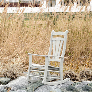 Pale gray poly rocking chair among wind blown dune grasses with a white fence behind it