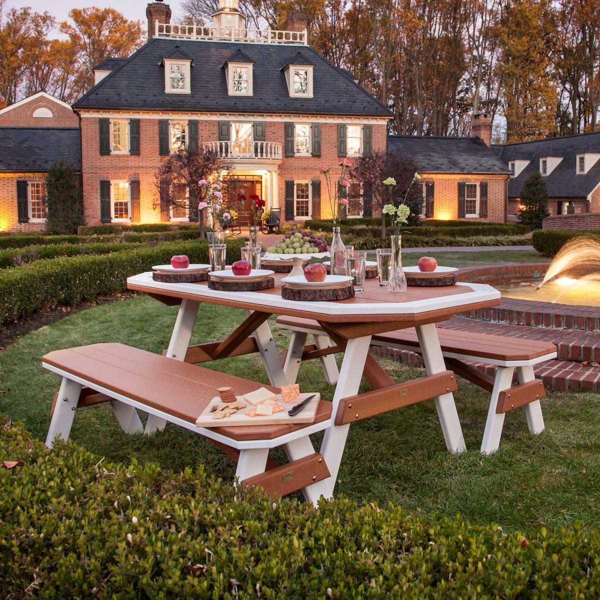 Cedar and white poly garden table with four benches set with apples and cheese on a lawn before a brick estate