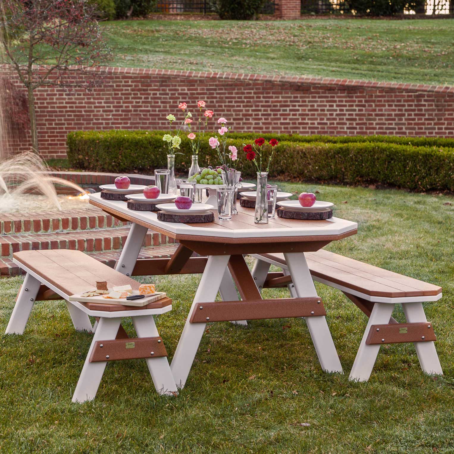 Cedar and white poly garden table with four benches set with apples and cheese on a lawn before a brick estate