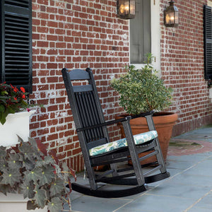 Black poly rocking chair with tropical print seat cushion on a bluestone porch beside begonias and a brick wall