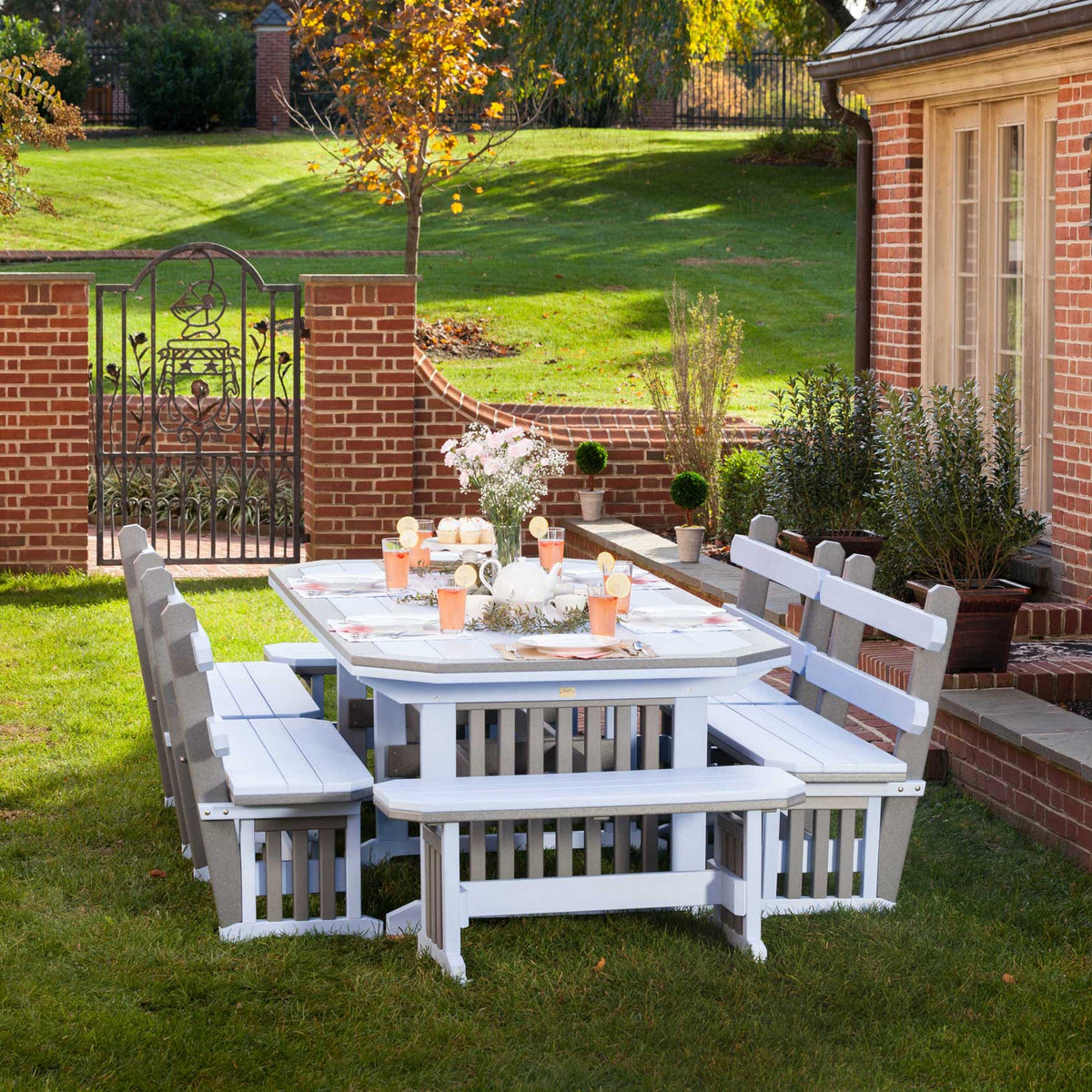 White and weatherwood Mission poly table with four benches set for a party on a lawn by a brick garden gate