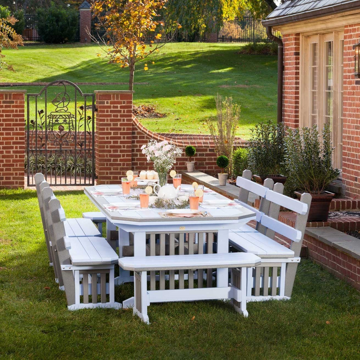 White and driftwood gray poly patio dining table with four slatted benches set for a meal on a lawn beside a brick garden wall