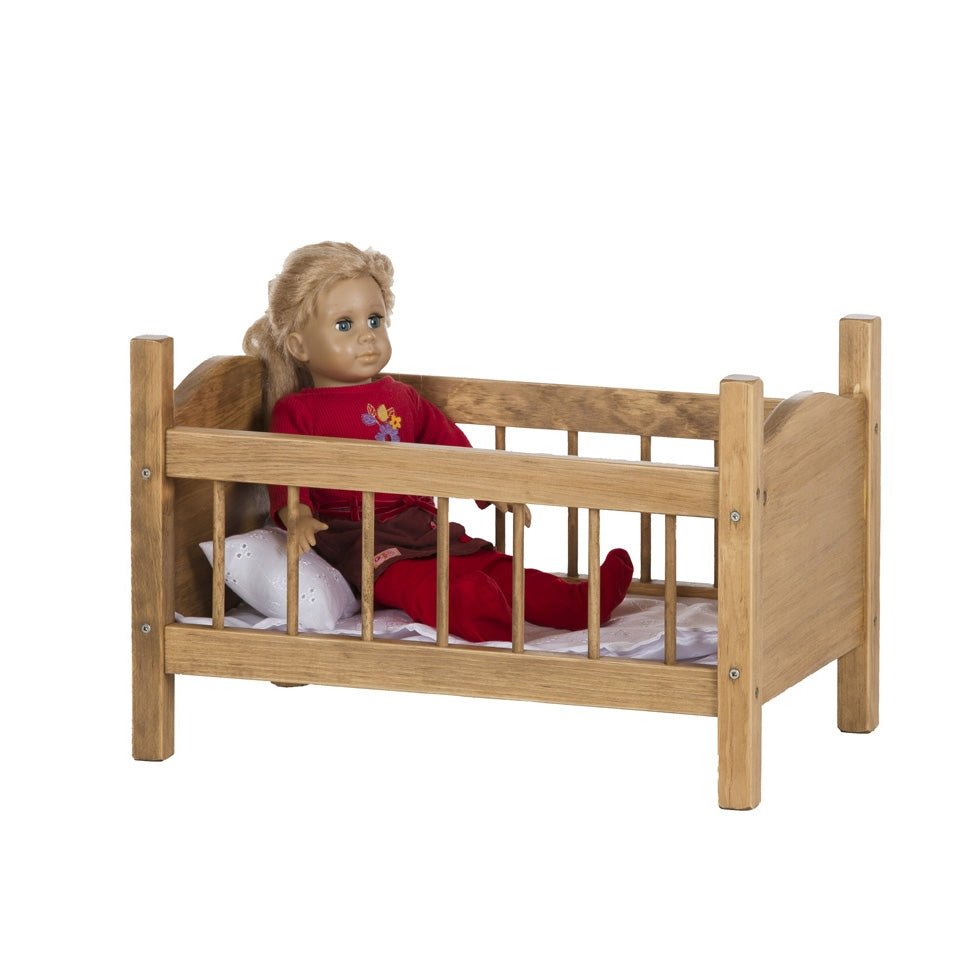 Wooden doll crib in a golden stain holding a blonde doll in a red outfit against a white pillow and sheet
