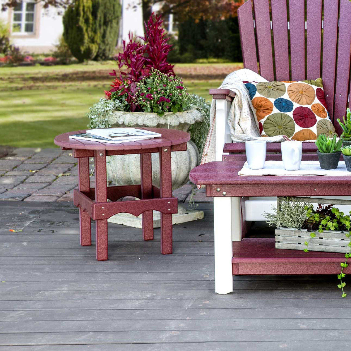 Cherrywood red poly round side table with scalloped skirt on a gray deck next to a matching glider and planter