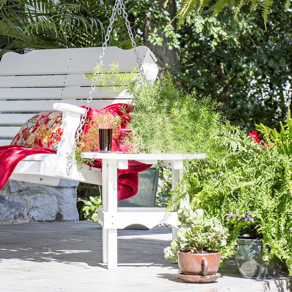 White poly round side table beside a hanging porch swing with a red throw, surrounded by ferns and potted plants