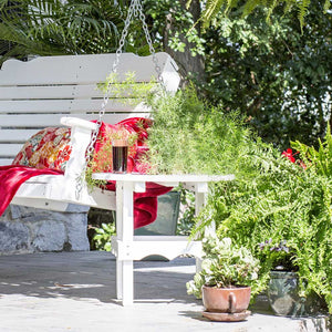White poly round side table beside a hanging porch swing with a red throw, surrounded by ferns and potted plants