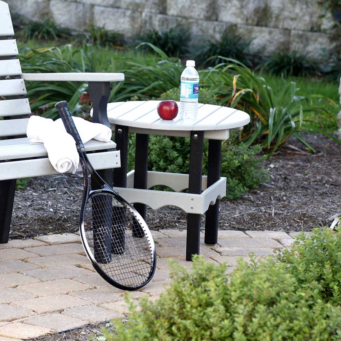 Two tone gray and black round poly patio side table on pavers next to a bench, with towels and a tennis racket