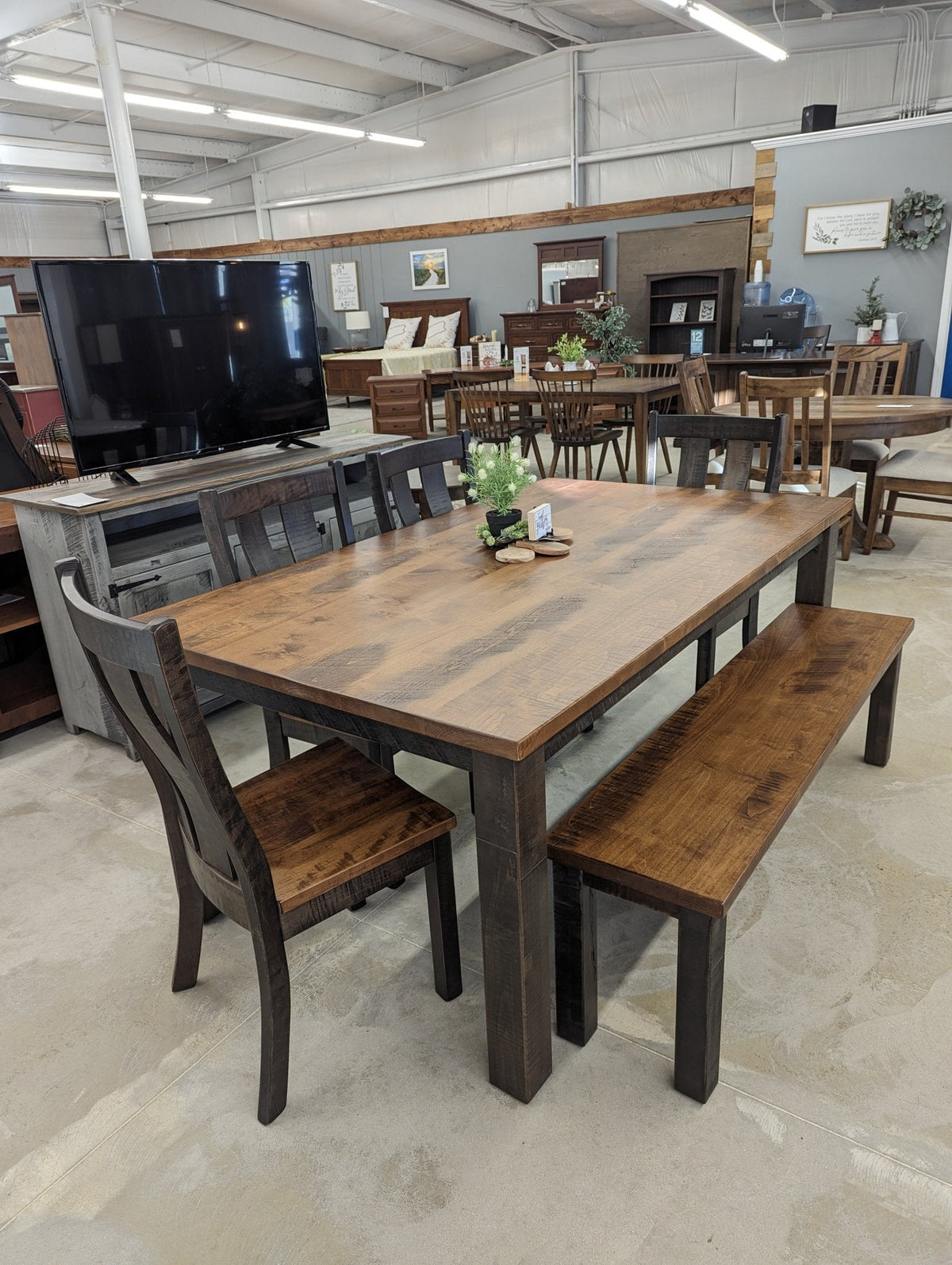 Reclaimed wood farmhouse table with dark distressed legs, matching slat back chairs and bench, displayed in a furniture showroom