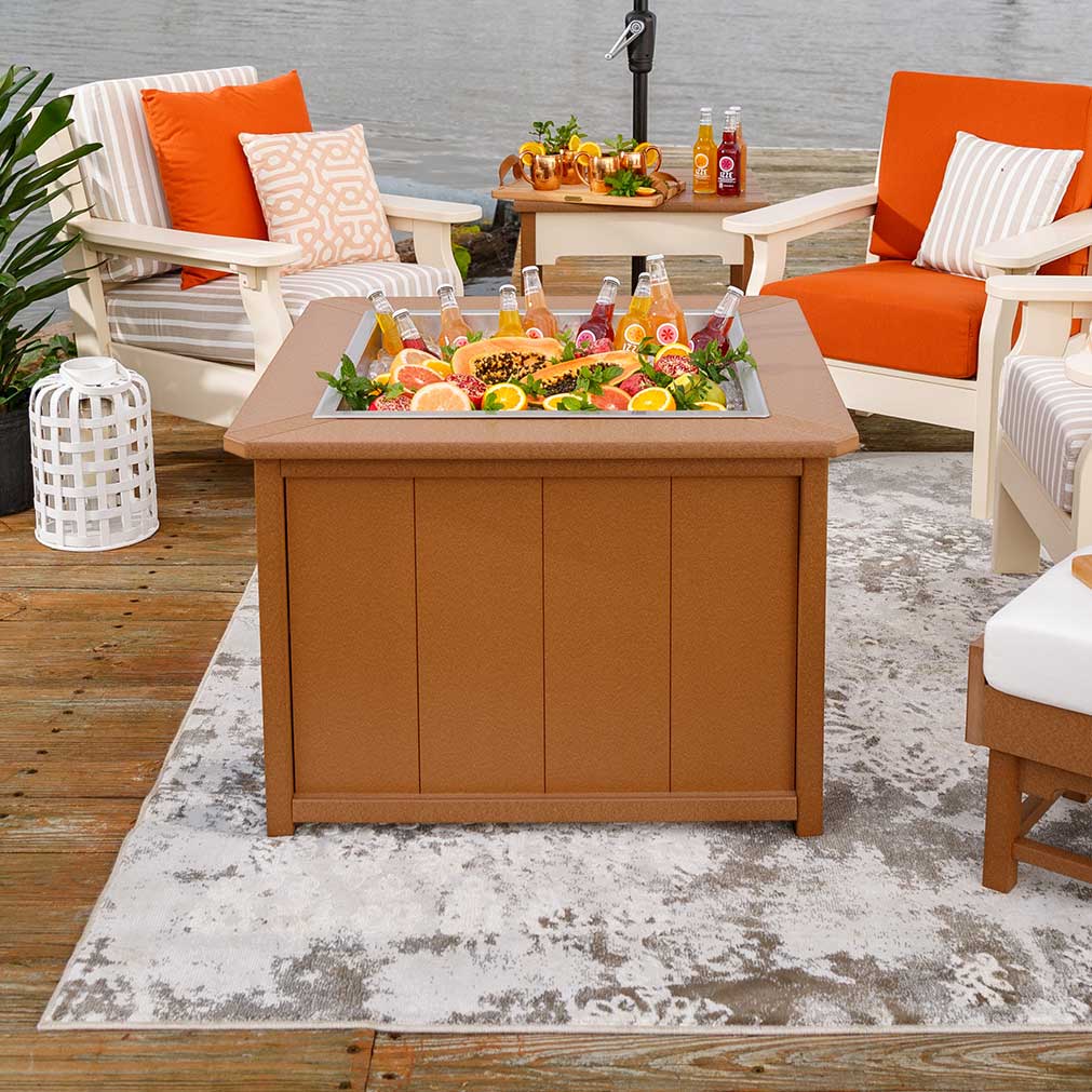 Cedar poly ice pit table filled with iced bottles and fruit, set on a rug between white deep-seating patio chairs