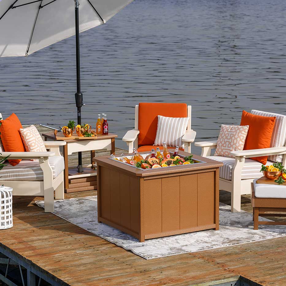 Cedar poly ice pit table stocked with drinks on a lakeside dock beside white cushioned patio chairs and an umbrella