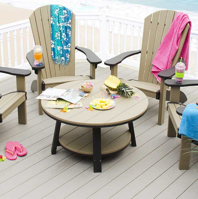 Round weathered wood and black poly patio coffee table with lower shelf, surrounded by Adirondack chairs on a beach deck