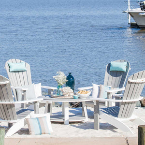 Round gray poly coffee table set with drinks between four white and gray Adirondack chairs at the water's edge