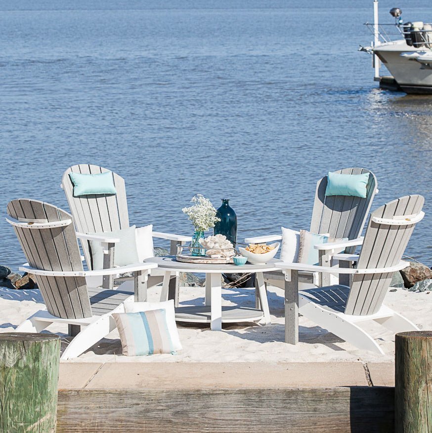 Four gray and white poly Adirondack chairs circled around a round coffee table with snacks on a waterfront beach