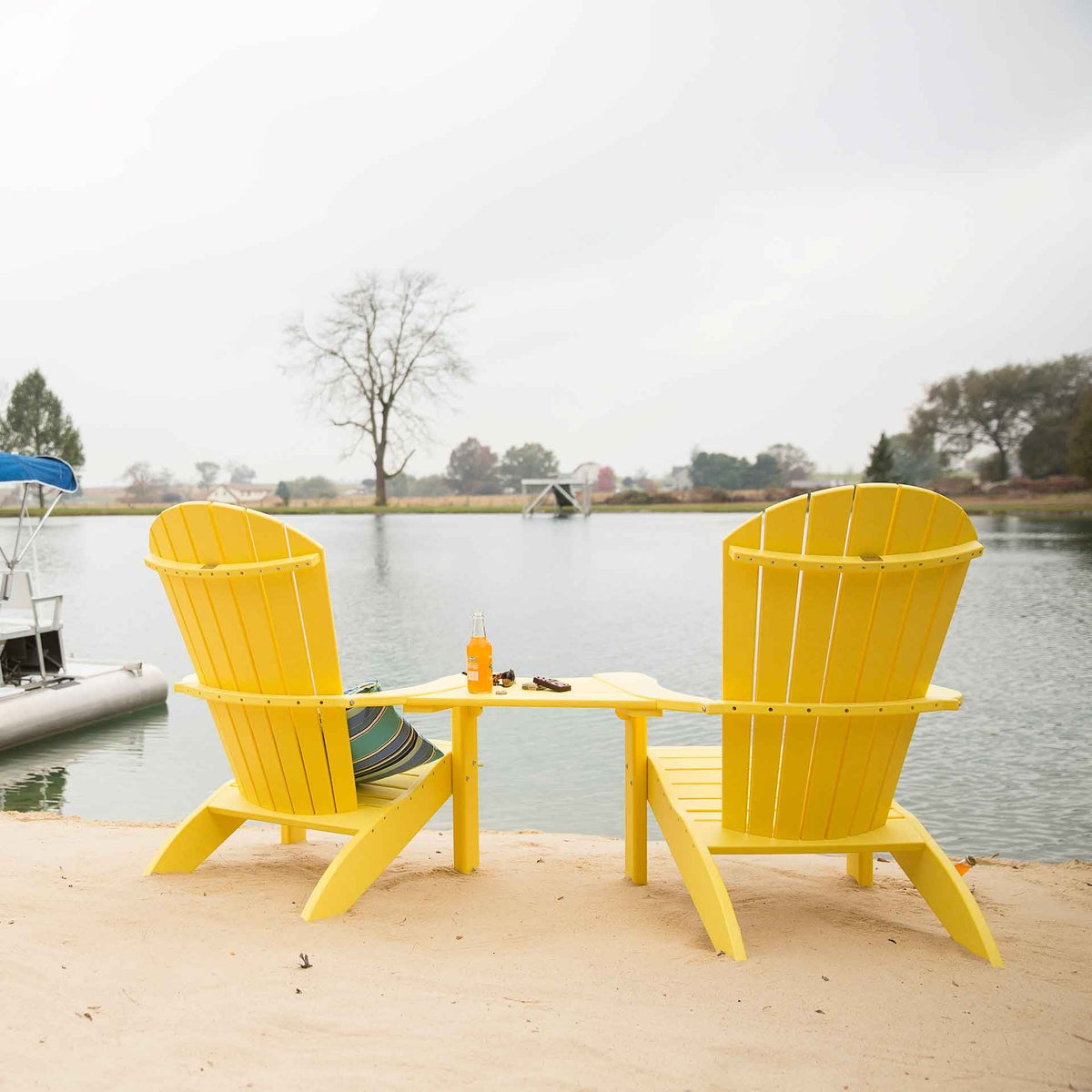 Pair of yellow Adirondack chairs with a joining table facing a foggy lake from a sandy shoreline