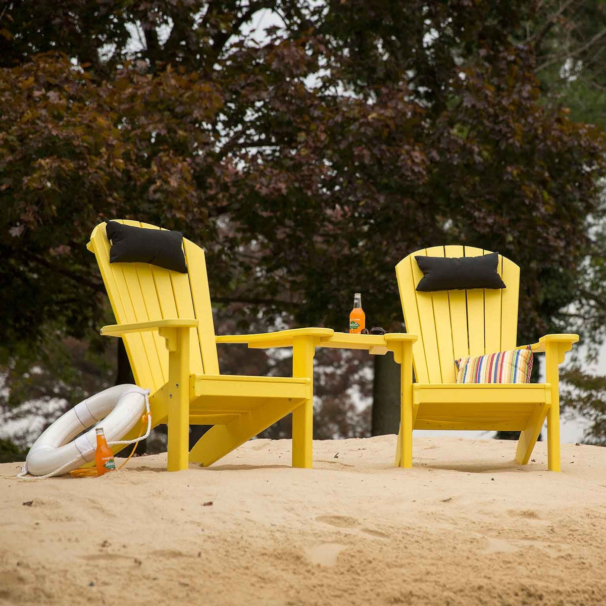 Two yellow poly Adirondack chairs joined by a curved center table with black headrest pillows on a sandy beach