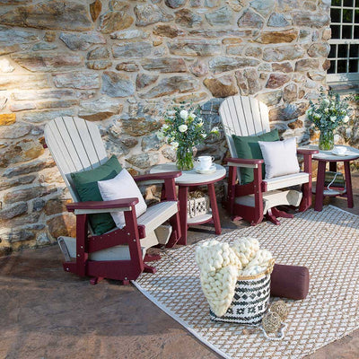 Pair of weatherwood and cherry Adirondack gliders with green pillows and round side tables on a stone patio